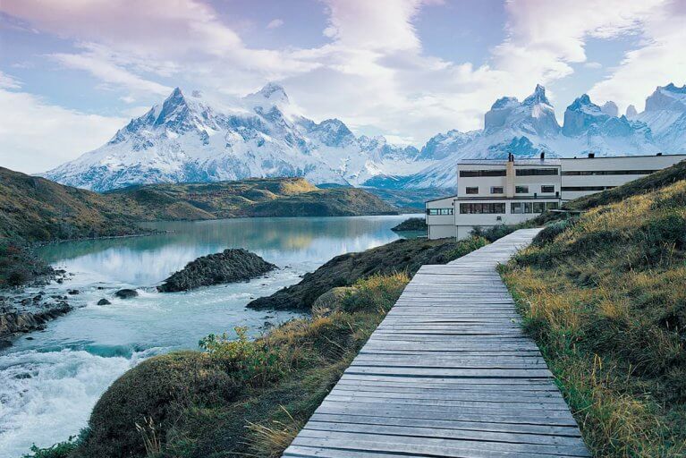 A wooden path along light blue water leads to luxury Explora Hotel with mountains in the distance