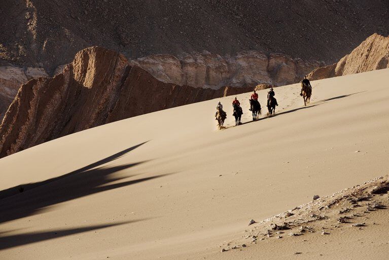 Five people horseback riding on a sand dune in the Atacama Desert in front of mountains