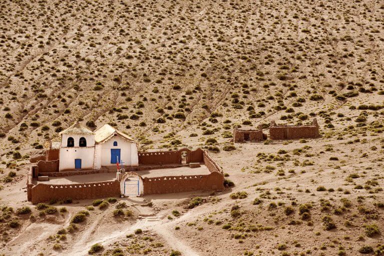 A small, isolated white church with blue doors between hills in Atacama Desert during luxury trip to Chile