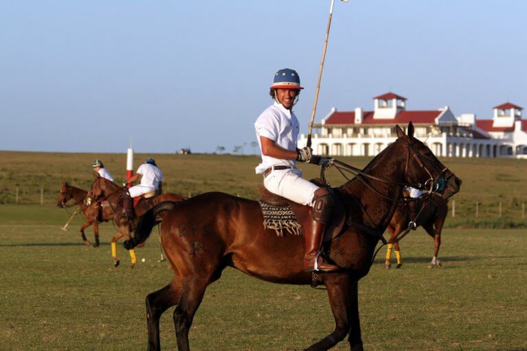 Men playing polo on brown horses with Estancia Vik in background in Uruguay