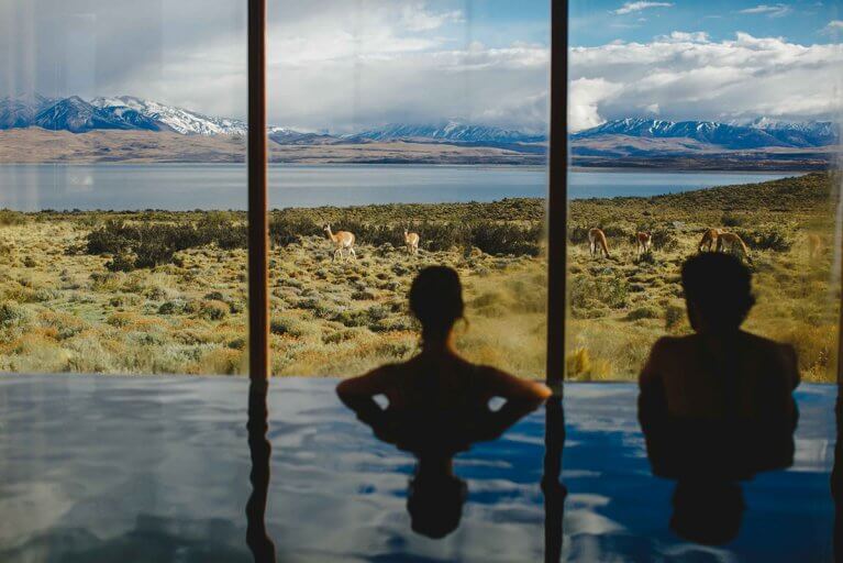 Two people enjoying the indoor pool at the spa of Tierra Patagonia with a view of mountains, a lake, and guanacos