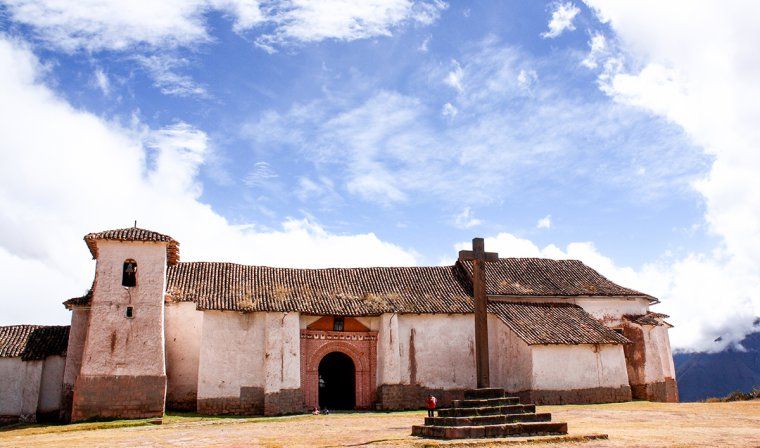 stone cross in front of a white church with adobe tile roof in small town in Peru's Sacred Valley
