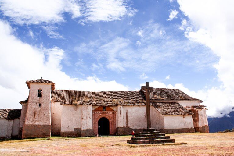 stone cross in front of a white church with adobe tile roof in small town in Peru's Sacred Valley