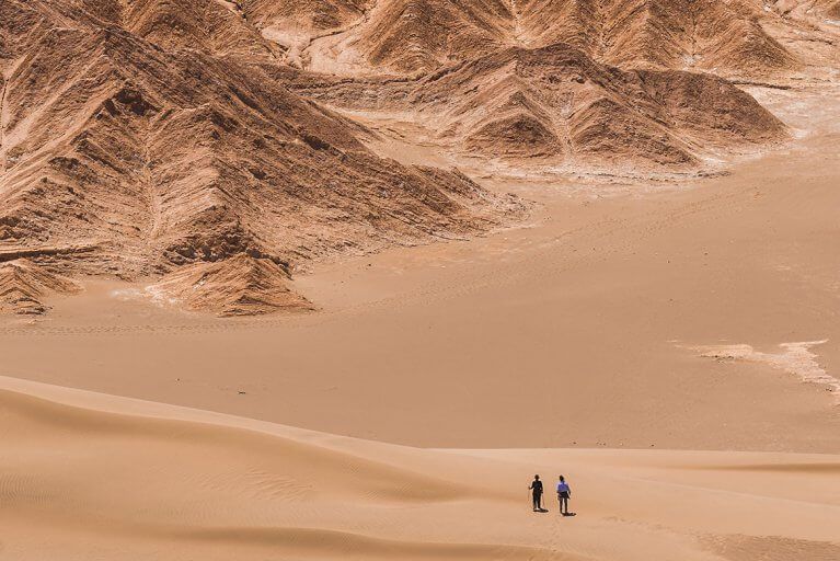 Two people seen from a distance hiking down reddish sand dunes on a private Atacama Desert tour