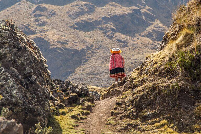 Back of a traditionally dressed Peruvian woman standing on a dirt path looking toward mountains