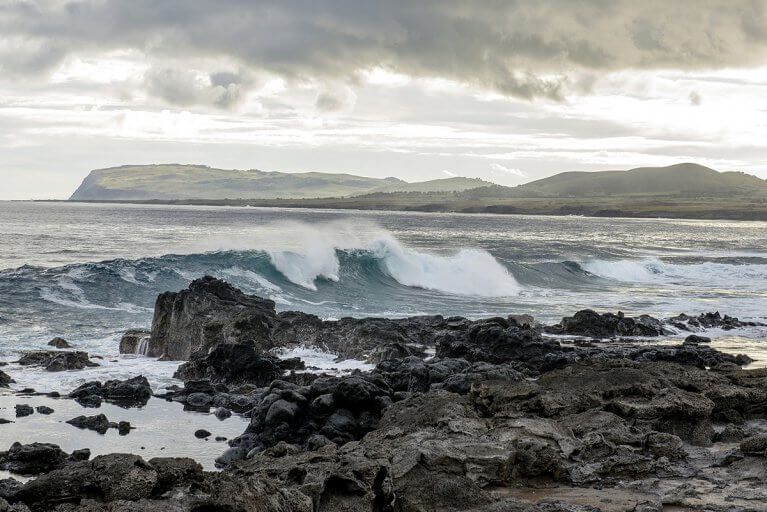 Waves crash on a rocky beach with more of the coast of Easter Island in the distance