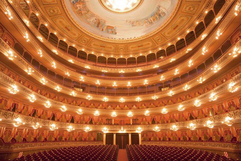 Red empty chairs and golden ornate interior of Teatro Colon with lights on during a private tour