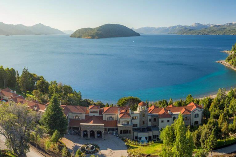 Villa-style hotel called Villa Beluno with red roofs surrounded by forest on the shore of a lake with mountains in the distance