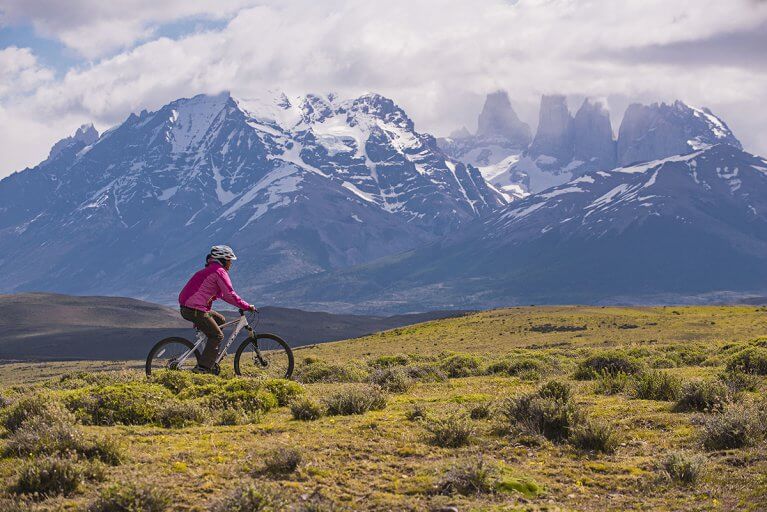 A woman biking through a natural field on a private Patagonia tour, with snowy mountains in the distance