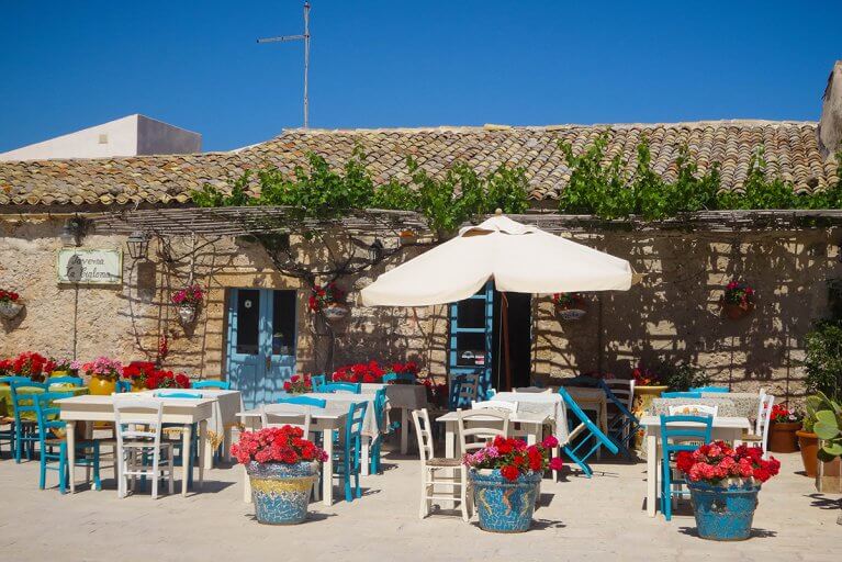 Charming cafe with outdoor tables and red flowers in fishing village Marzamemi