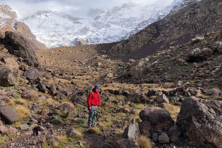 Man in a red jacket during a hiking excursion in Toubkal National Park, with snowy peak behind him
