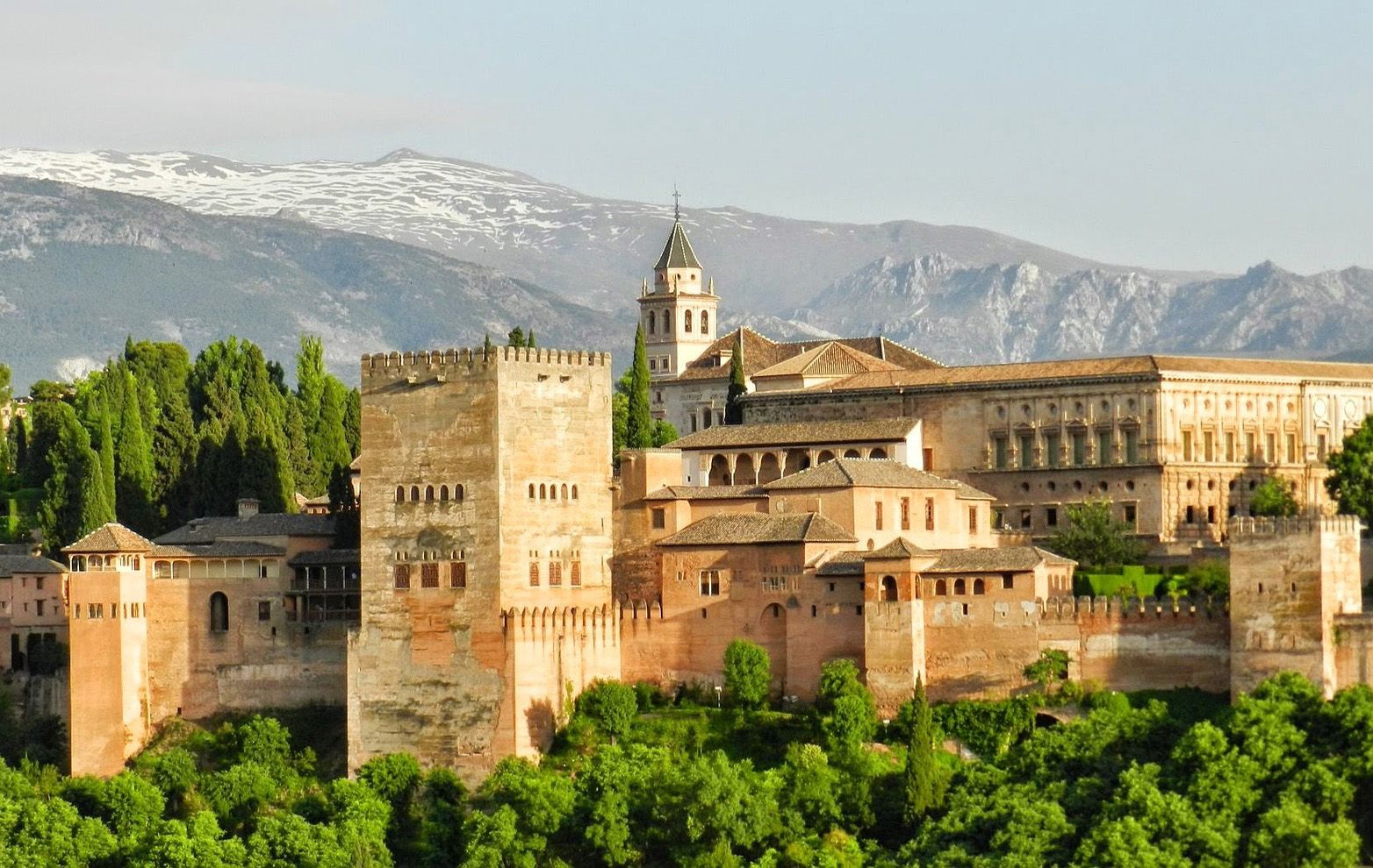 Alhambra sits on a hill surrounded by trees with mountain backdrop in Granada, Spain
