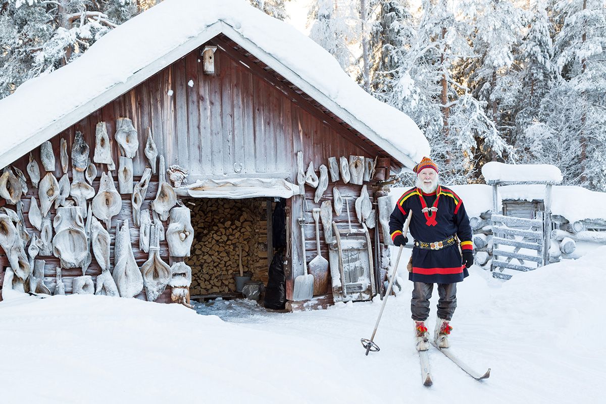 Sami man on skis smiling in front of a cabin filled with firewood during experiential travel to Swedish Lapland