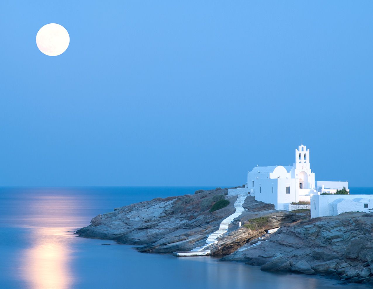 White Chrysopigi church sits on a rocky peninsula at night below a full moon