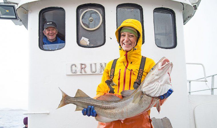 Woman smiling with an arctic cod as the captain looks on, during a fishing excursion in Lofoten