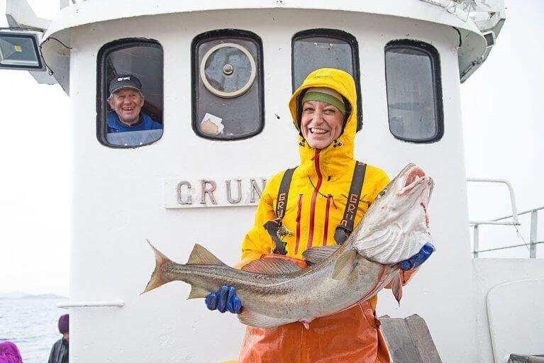 Woman smiling with an arctic cod as the captain looks on, during a fishing excursion in Lofoten