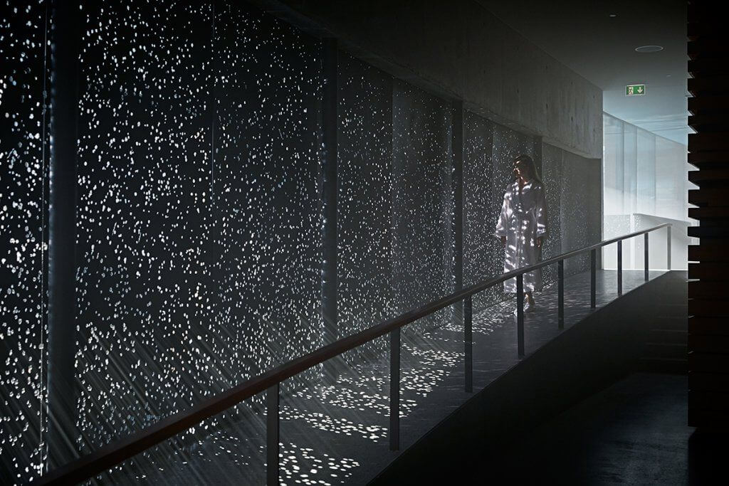 Woman in a bathrobe walking through a corridor with light beams shining through holes in the wall