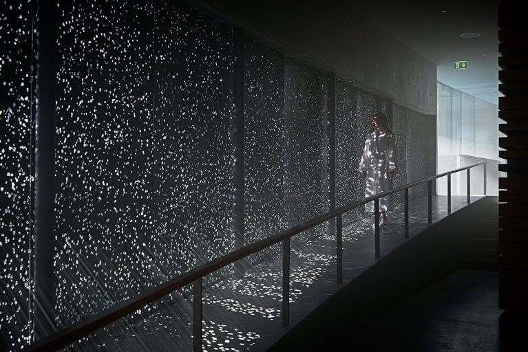Woman in a bathrobe walking through a corridor with light beams shining through holes in the wall