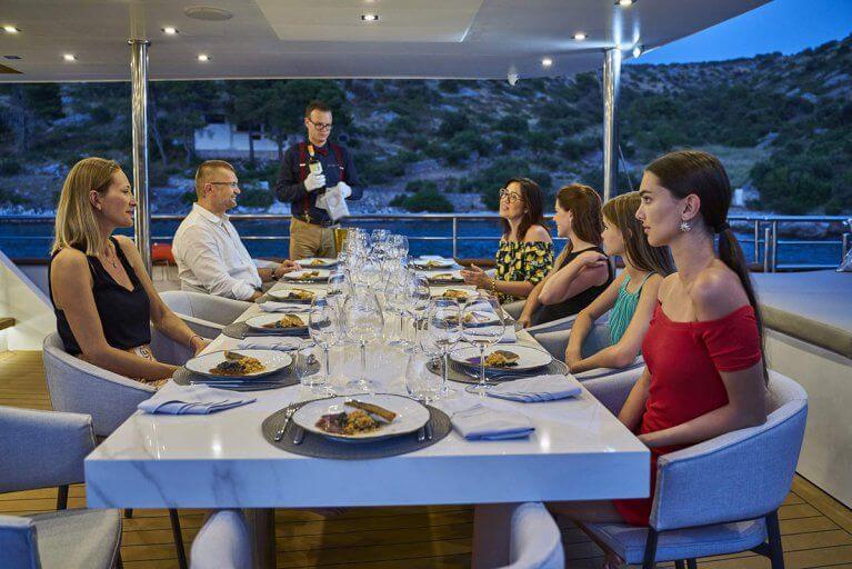 People sit at a long dinner table on aft deck of a private yacht sailing in Adriatic sea