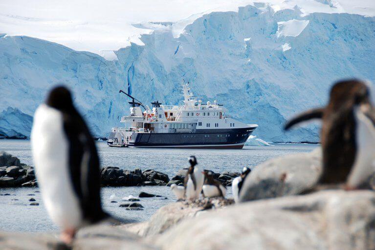 Penguins playing on rocks in Antarctica with the Hanse Explorer yacht cruising in the background
