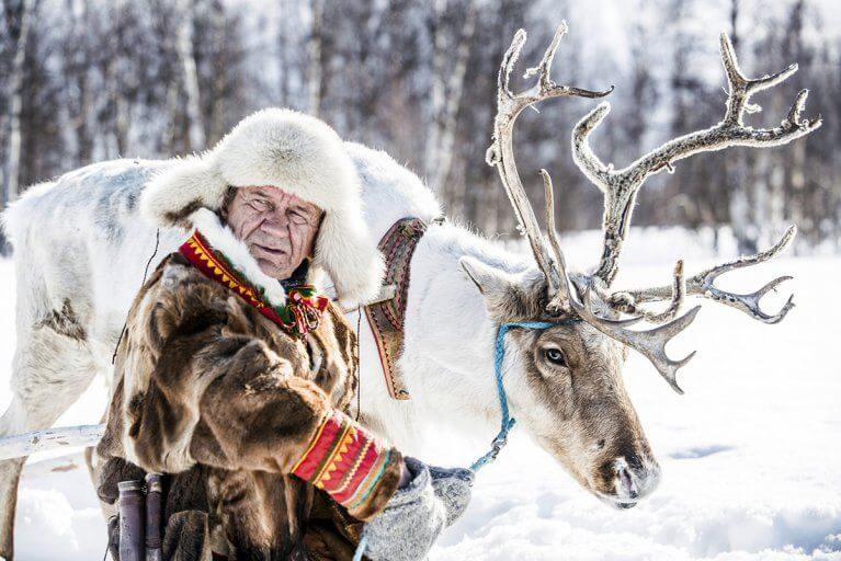 Sami man dressed in furs leading a reindeer through a snowy forest