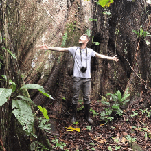 Man with outstretched arms in front of massive tree in rainforest