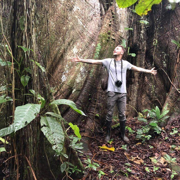 Man with outstretched arms in front of massive tree in rainforest